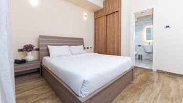 Un dormitorio moderno con estructura de cama de madera, una mesita de noche con un florero y un baño visible a través de una puerta abierta.