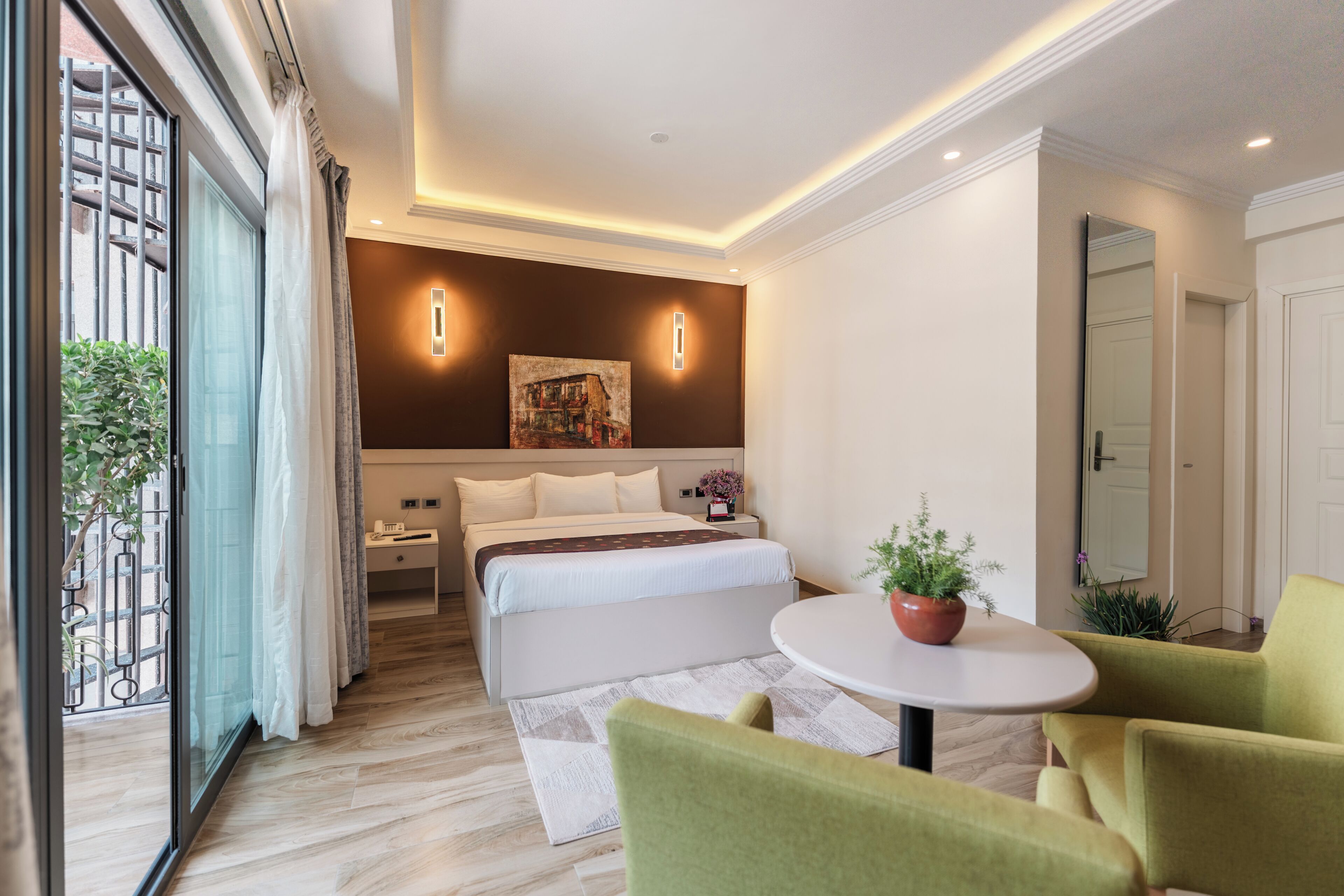 A modern hotel room with a large bed, a small table with a plant, and a view of the outdoors through a sliding door.