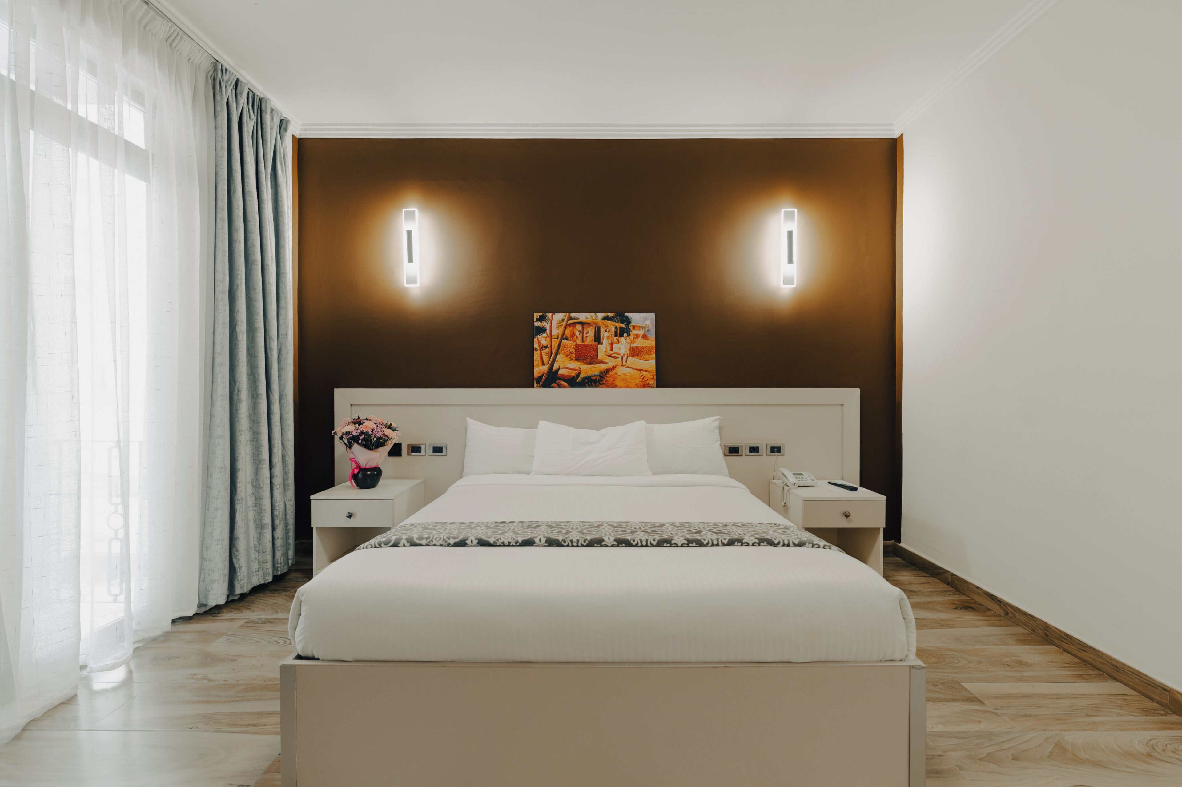 A modern hotel room with a large bed, bedside tables, a nightstand, a painting on the wall, and a window with curtains.