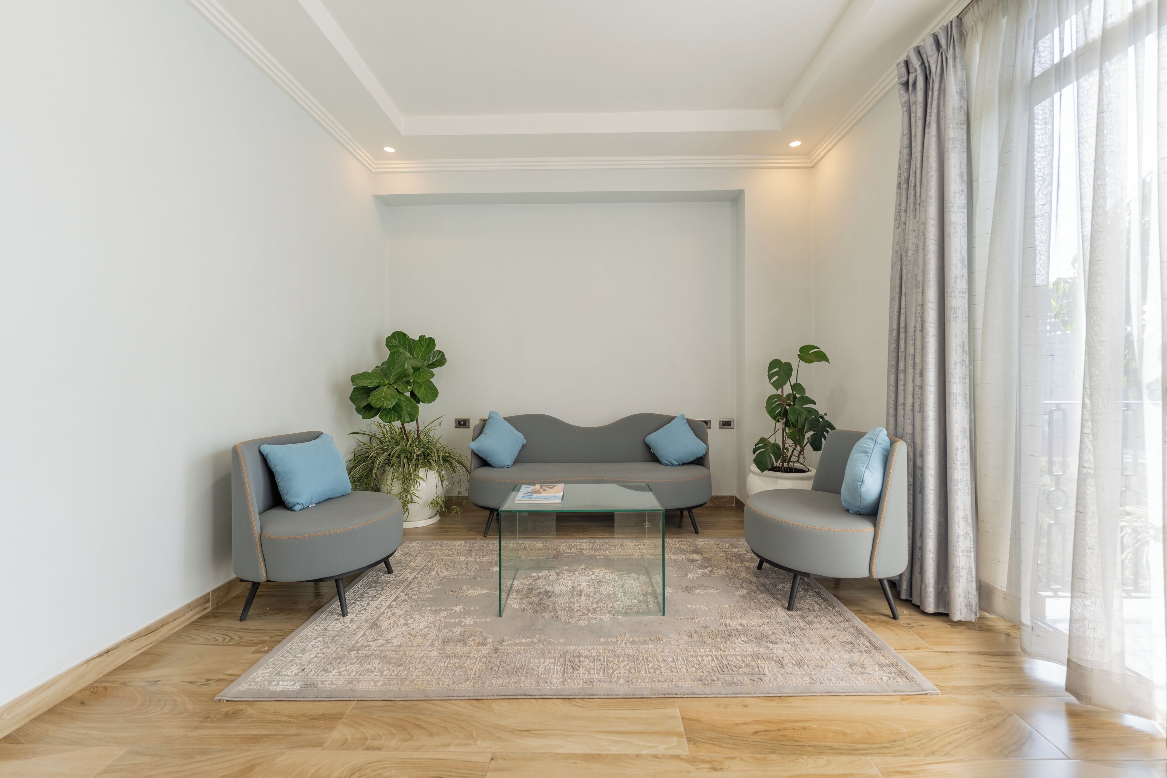 A modern living room with a grey sofa set, a glass coffee table, and large windows with curtains.