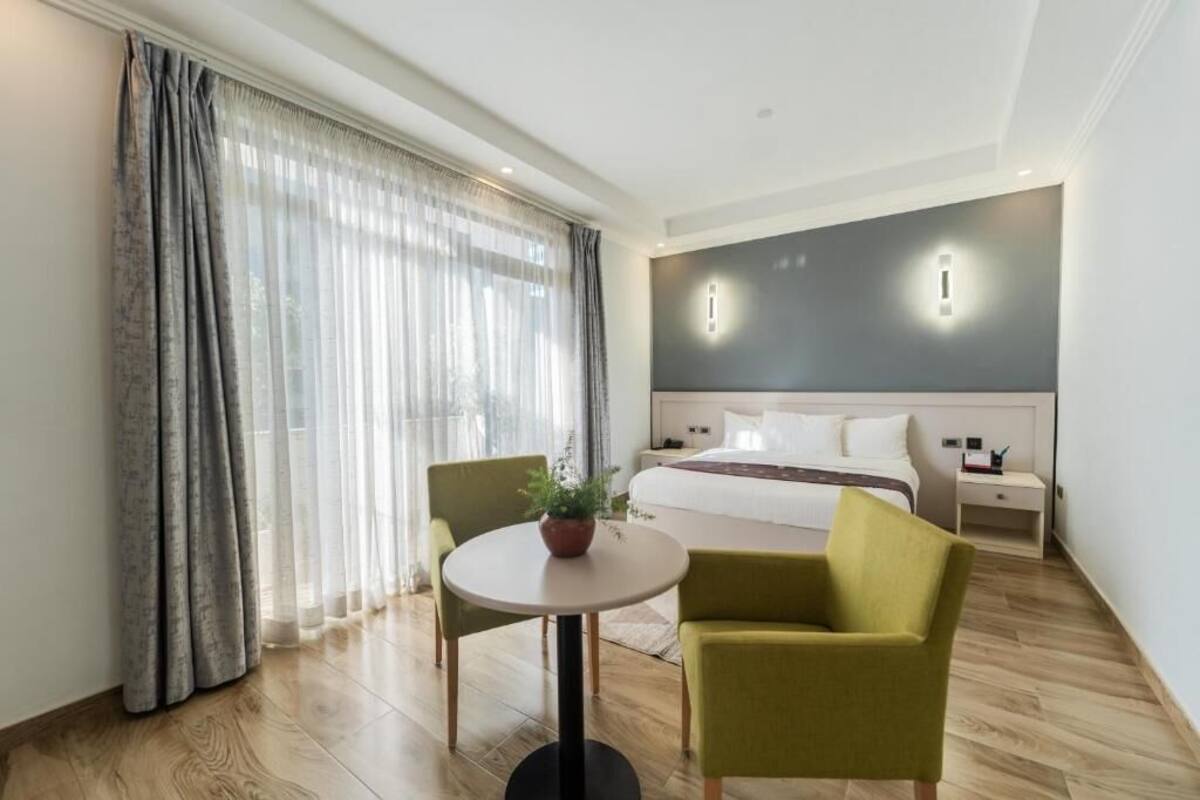 A hotel room with a bed, a bedside table, a nightstand, a round table with a chair, and a potted plant.