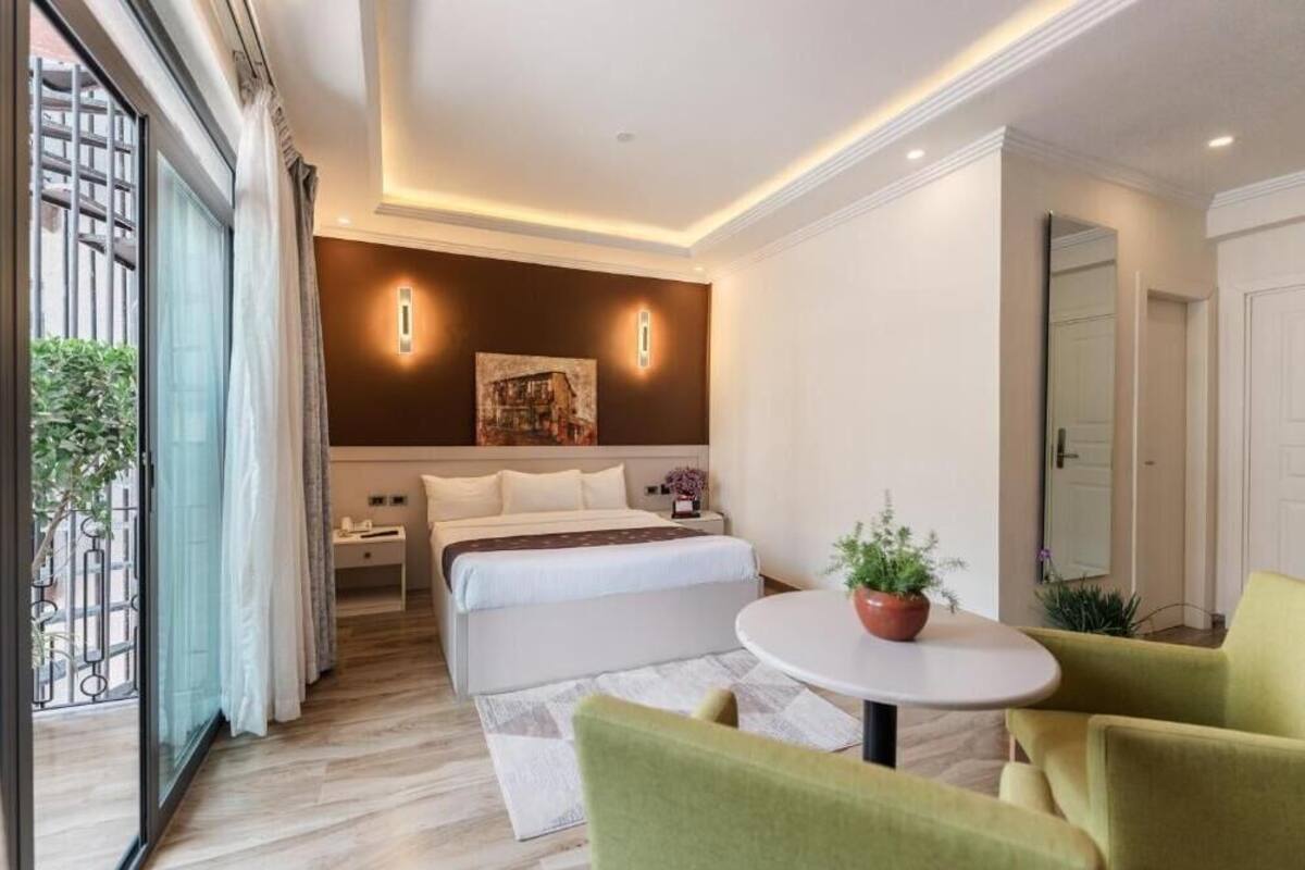 A hotel room with a bed, bedside table, lamp, potted plant, and a view through a window.