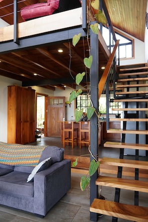 A spacious loft-style room with a high ceiling, a wooden staircase, and a large window providing natural light.