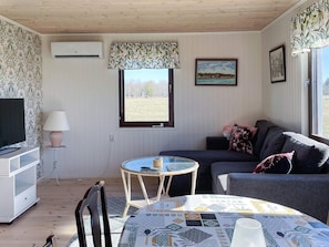 Ein Zimmer mit Fernseher, einem Tisch mit gemustertem Tischläufer, einer Couch und einem Fenster mit Blick ins Freie, sowie einer Klimaanlage.