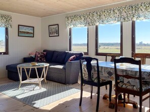 Una sala de estar acogedora con un sofá, una mesa redonda y sillas, ventanas amplias con vistas al exterior, y dos cuadros en la pared.