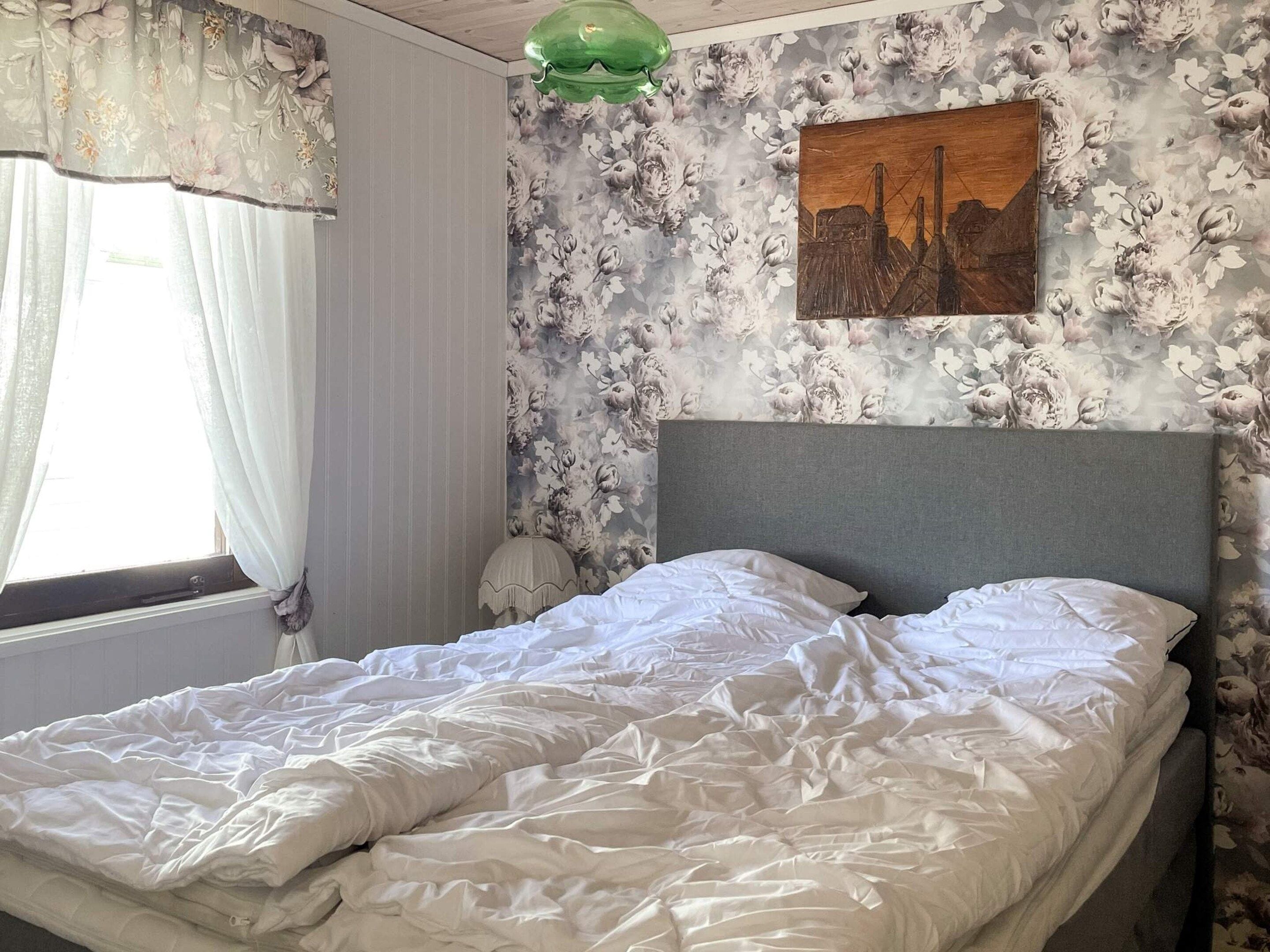 Ein Schlafzimmer mit einem Bett, floralem Wandpapier, einem Gemälde und einem Fenster mit Vorhängen.