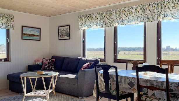 Un salon chaleureux avec un canapé, une petite table ronde et une table à manger avec des chaises. Il y a deux fenêtres avec des rideaux, et un tableau est visible au mur.