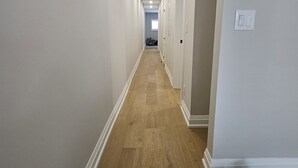 Kapea käytävä, jossa on puulattia ja valkoiset seinät, johtaa huoneeseen, jossa on ovi.