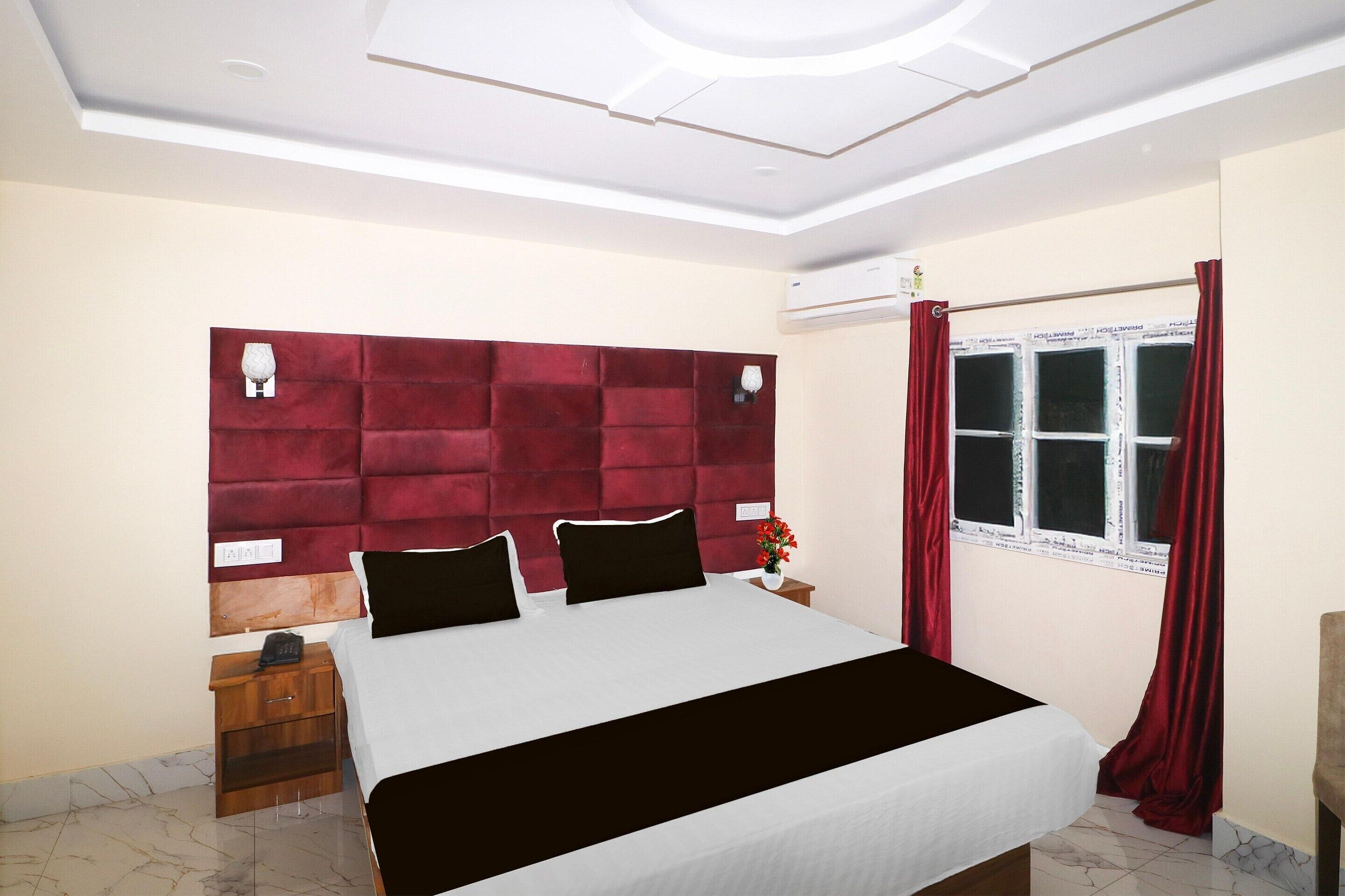Une chambre avec un grand lit, une table de chevet en bois, une tête de lit rouge, une fenêtre avec des rideaux et un luminaire au plafond.