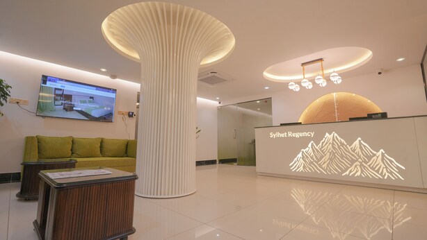 A reception area with a modern design, including a large pillar, a reception desk, a green sofa, and a wall-mounted TV. - Sylhet Regency Ltd. (Sylhet)