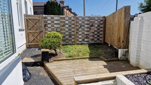 Un patio trasero con una terraza de madera, un arbusto pequeño y una cerca.