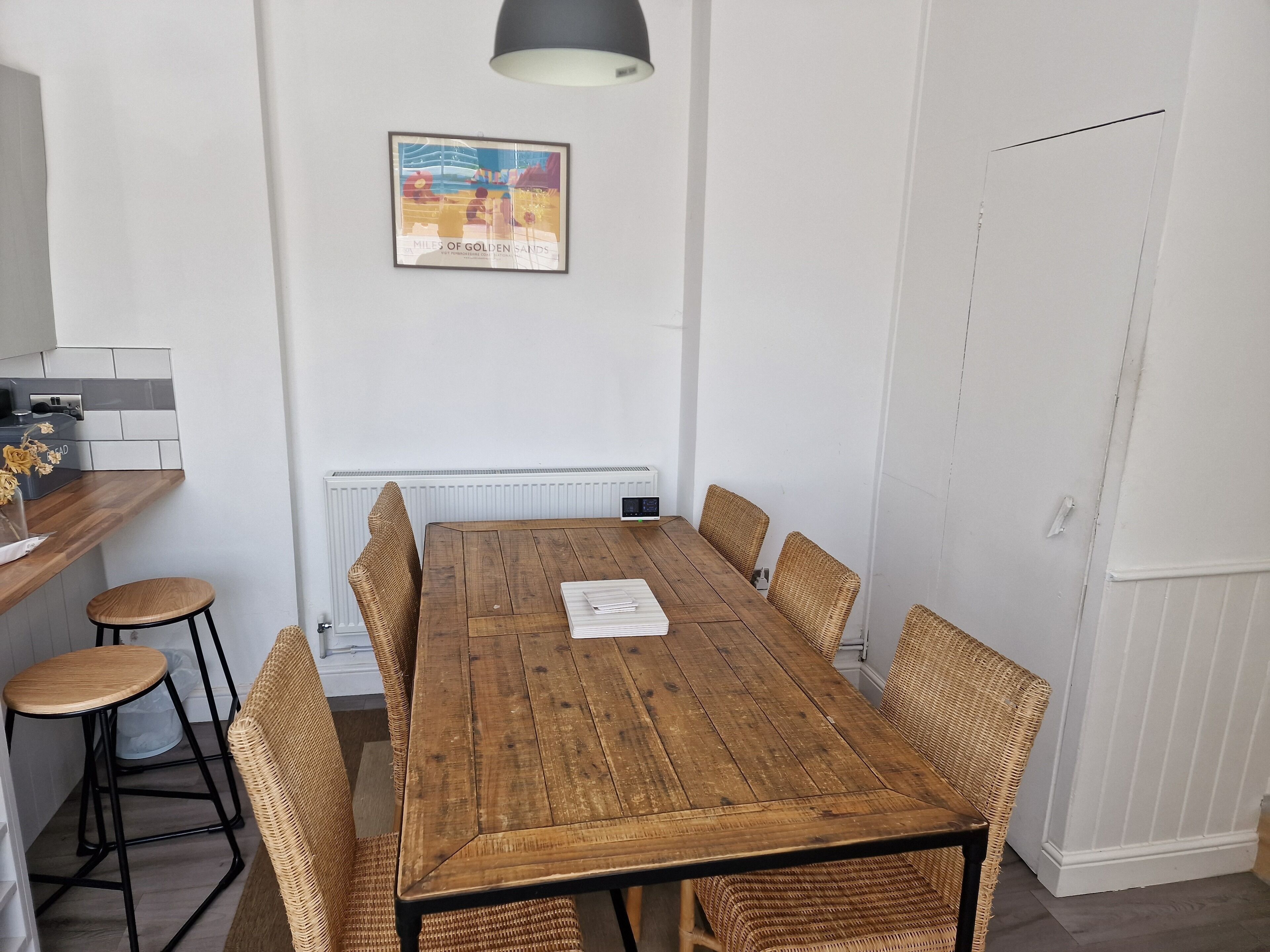 Un comedor con una mesa de madera, sillas de mimbre, un radiador blanco y un cuadro en la pared.