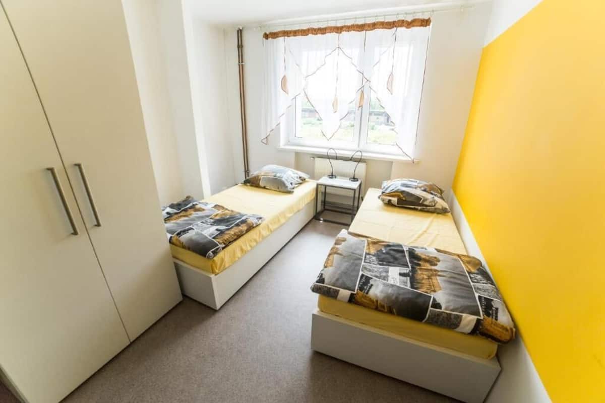 Una habitación pequeña y luminosa con dos camas, una ventana con cortinas y un armario blanco.