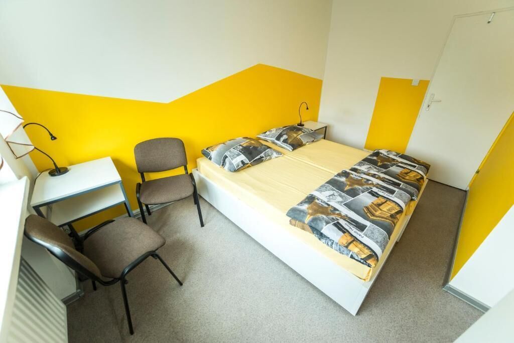A single bed with a patterned bedspread, a desk with a chair, and a lamp.