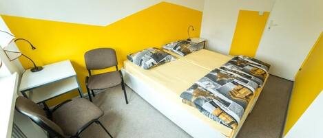 A single bed with a patterned bedspread, a desk with a chair, and a lamp.