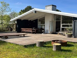 Área externa coberta com banco, mesa e cadeiras, cercada por um deck de madeira e um gramado.
