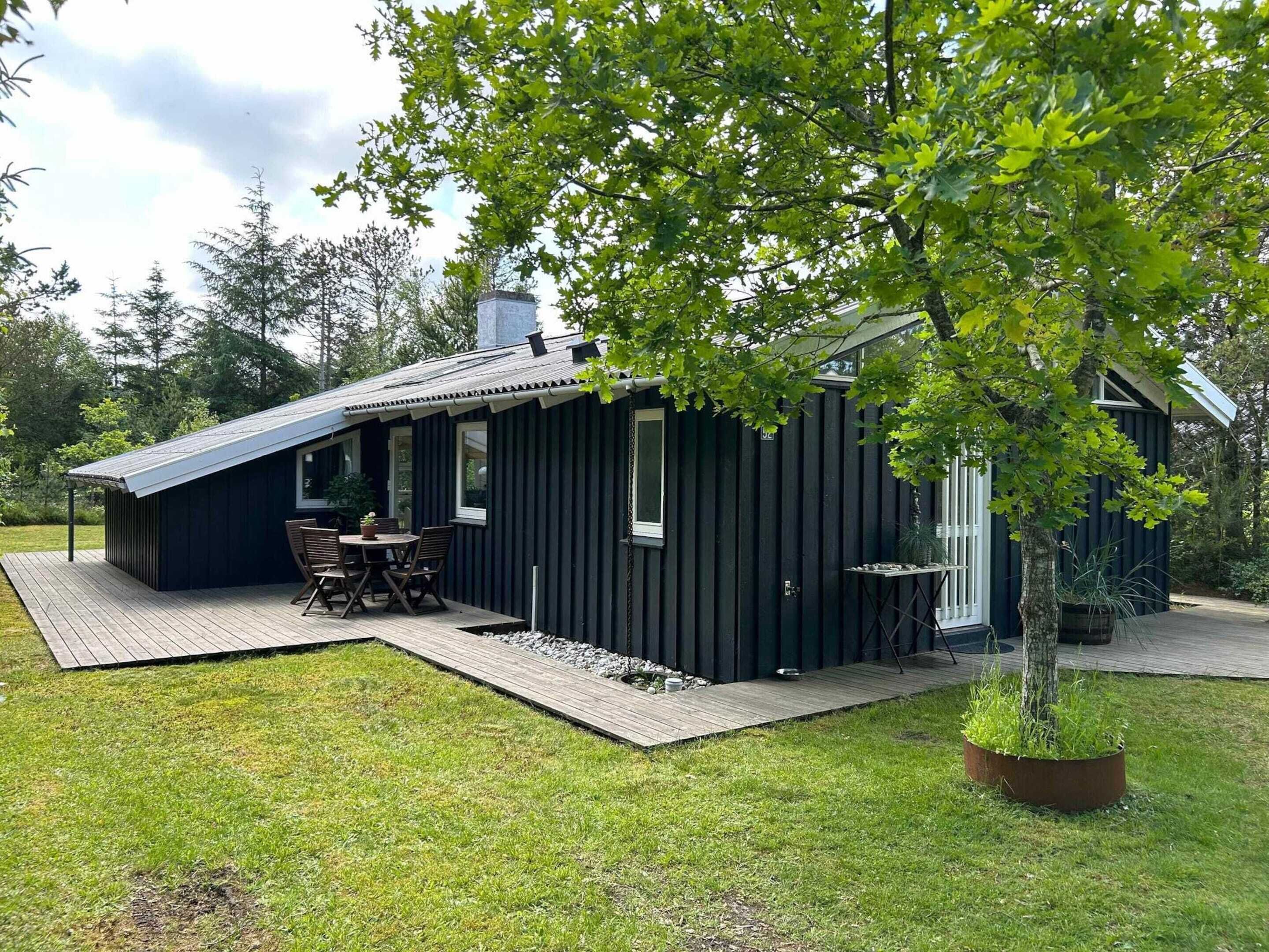 A small, dark-colored cabin with a sloped roof, a deck, and a dining area with outdoor furniture.