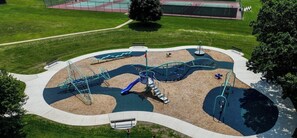 Vista aérea de un parque infantil con diversas estructuras de juego, una cancha de baloncesto y una zona verde circundante.
