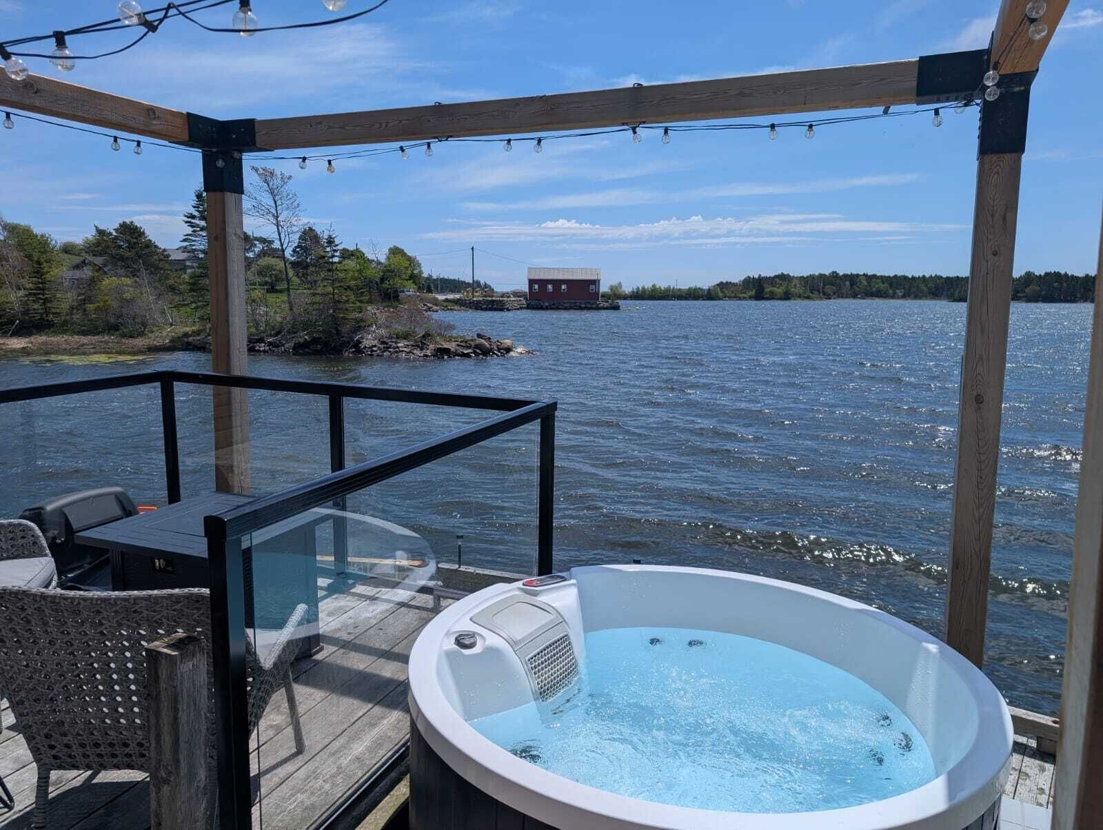 A hot tub on a wooden deck with a glass railing, overlooking a body of water and a distant building.