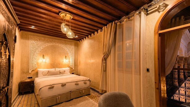 A bedroom with a bed, a chair, a nightstand, a decorative wall, and a view of an outdoor balcony. - Golden Riad (Rabat)