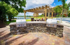 An outdoor grill area with stone walls and a pergola, surrounded by greenery and a view of a body of water. - Studio Near Disney & Universal - Westgate Lakes Resort (Orlando)
