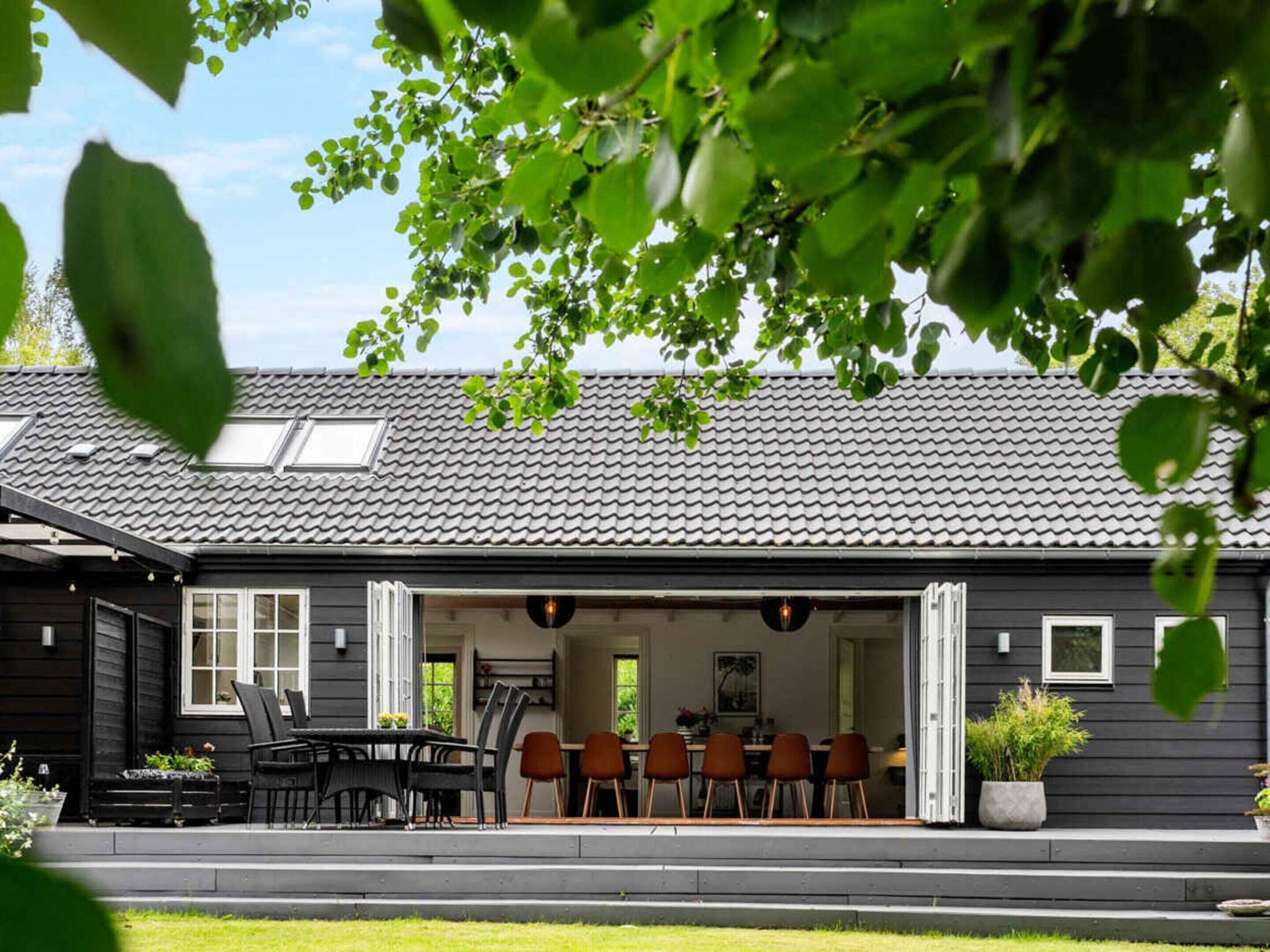 A modern house with a dark exterior, a tiled roof, and a covered outdoor seating area with chairs and a table.