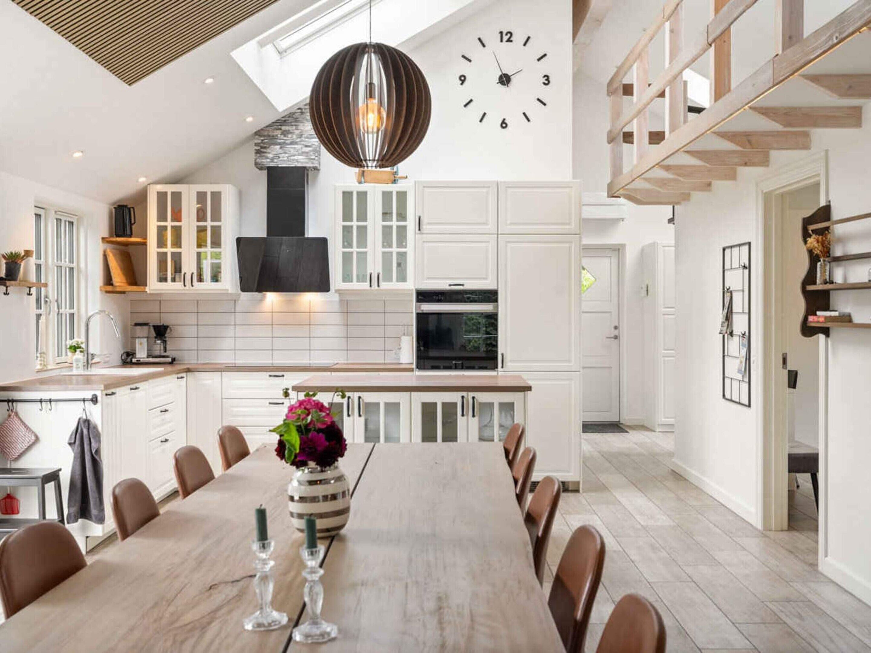 A modern kitchen with white cabinetry, a dining area with a long table, and a clock on the wall.