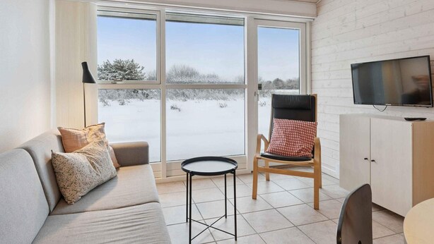 Ein modernes Wohnzimmer mit einer Couch, einem Sessel, einem kleinen Tisch, einem Fernseher und Blick auf eine verschneite Landschaft durch große Fenster.
