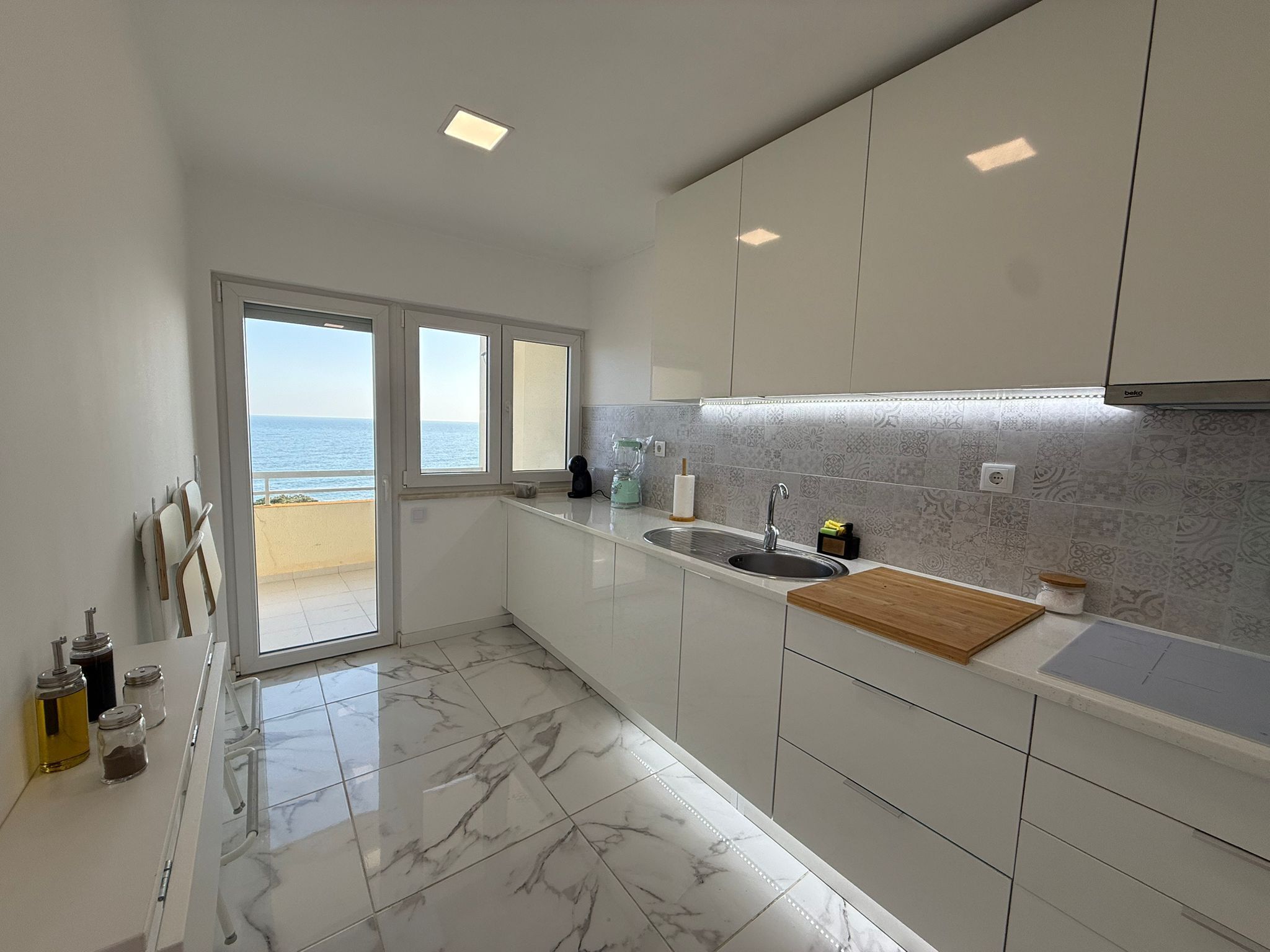 A modern kitchen with a view of the sea, white cabinets, a marble floor, and a wooden countertop.