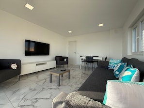 A modern living room with a flat-screen TV, a sofa, a coffee table, and a dining area with chairs.