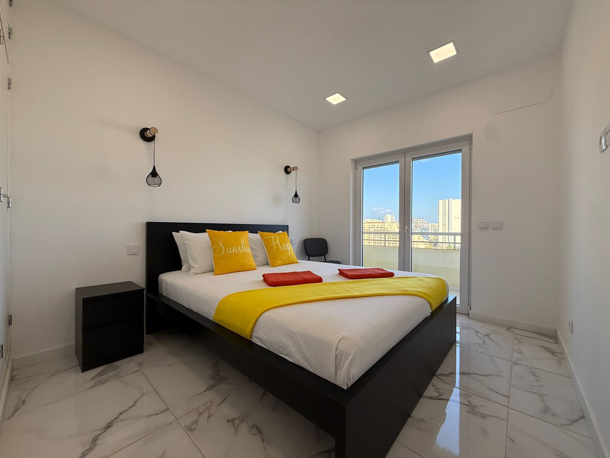 A modern bedroom with a large bed, a small bedside table, and a view of the city through a sliding door.