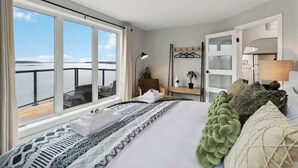 A hotel room with a large window, a bed with patterned bedding, a bedside table with a lamp, and a view of the sea. - Book 2 homes together. Ocean Views. Sleeps 8! (Lunenburg)