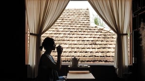 A person sitting on a tatami mat in a room with a wooden ceiling and a view of a tiled roof through a window with curtains.