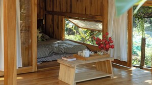 A wooden cabin with a bed, a small table with a vase, and a window overlooking greenery.