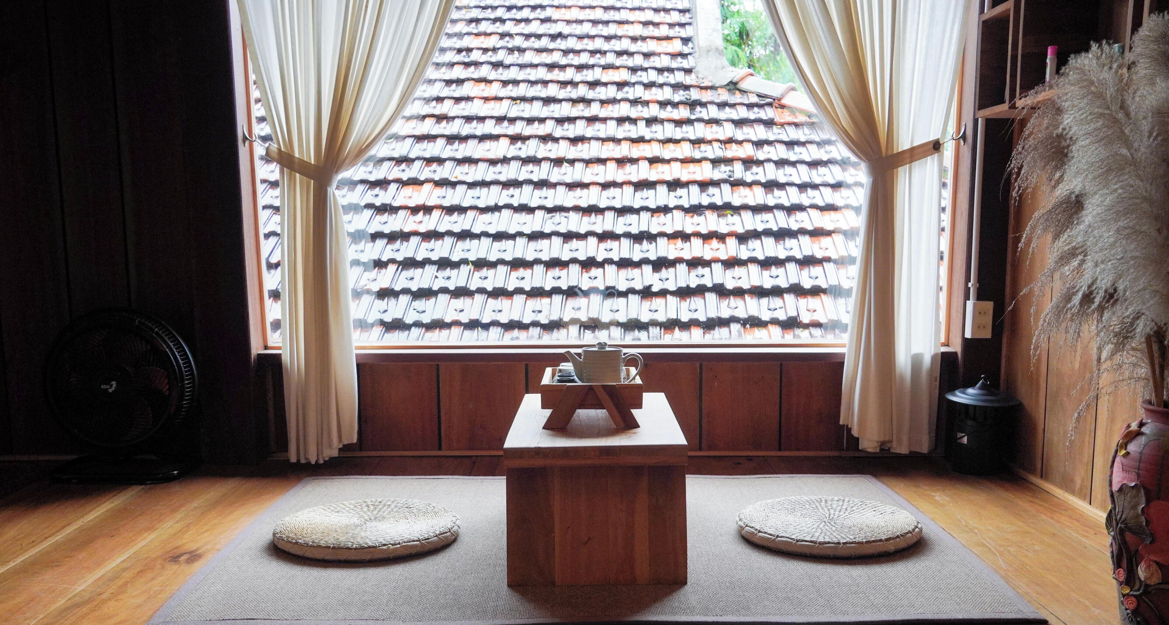Una habitación japonesa tradicional con tatamis, una mesa de madera baja y vista a un tejado de tejas por una ventana con cortinas.
