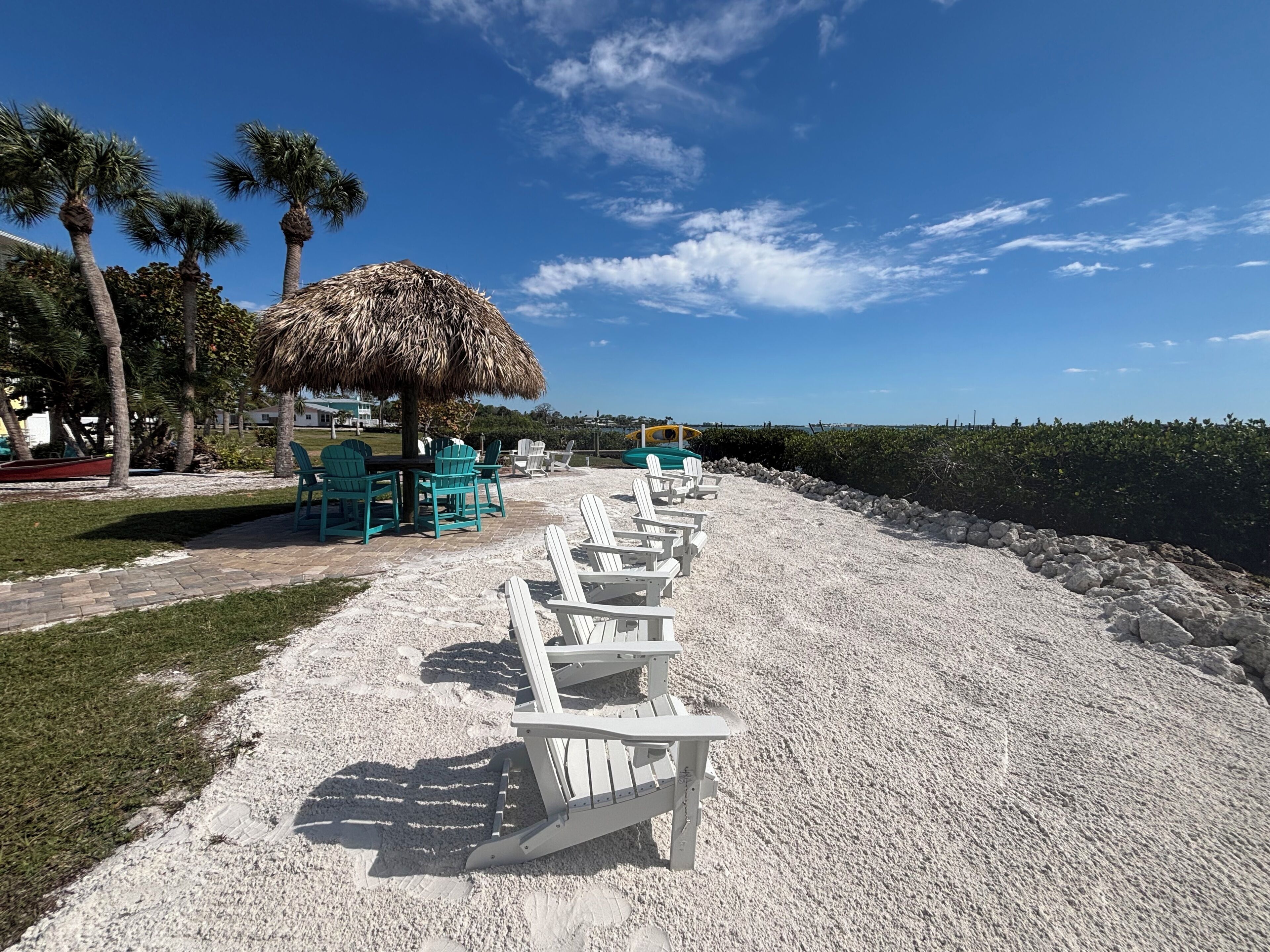 A beachside area with a row of white lounge chairs, a thatched umbrella, and a table with chairs.