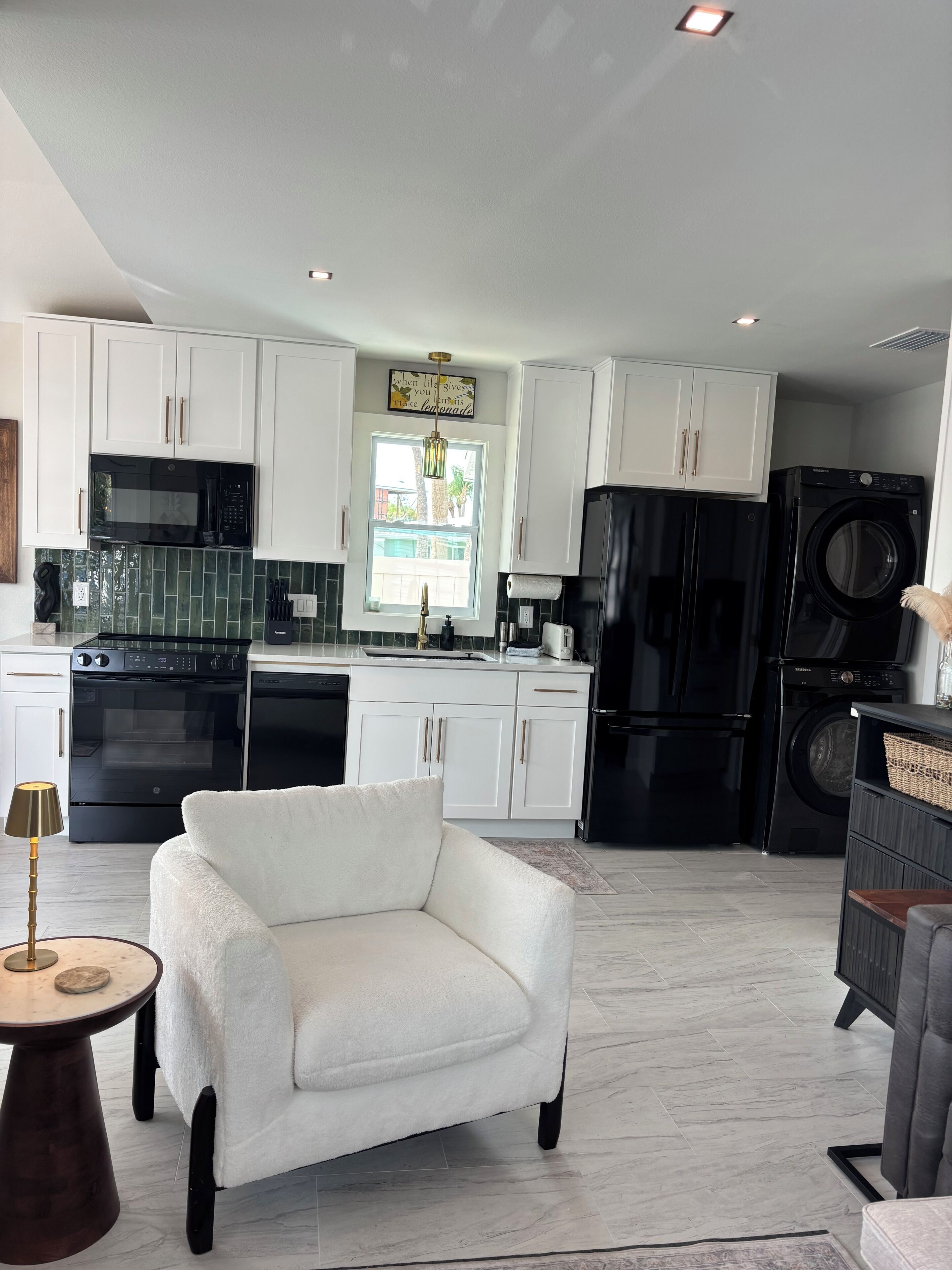 A modern kitchen with white cabinets, a black refrigerator, a washing machine, and a white armchair.
