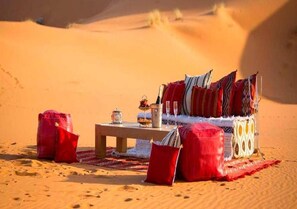 Wüstenlandschaft mit einem Sandhügel, einem Holztisch mit roter Decke, einer Sitzgelegenheit mit Kissen und einer kleinen Laterne.