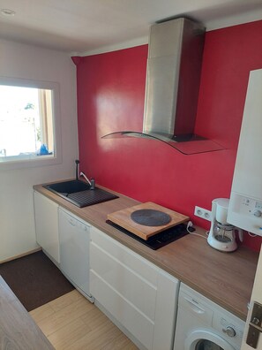 A compact kitchen with a red wall, a window, a dishwasher, a coffee maker, and a washing machine. - Gd Appartement 74 M2  1300 m plages (Le Pradet)