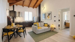 A modern living room with a sofa, dining table, and chairs, a kitchen area with cabinets and a stove, and a hallway leading to a bathroom.