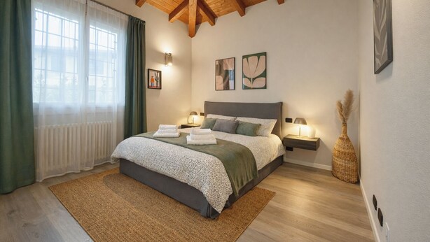 A bedroom with a wooden ceiling, a bed with a green and white bedspread, two bedside lamps, and a window with green curtains.