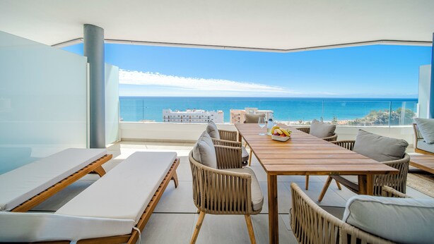 Een balkon met uitzicht op zee, een houten tafel, rieten stoelen en een bank.