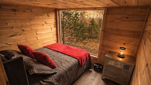 A cabin bedroom with a bed, pillows, a nightstand with a lamp, and a power tool on the floor.
