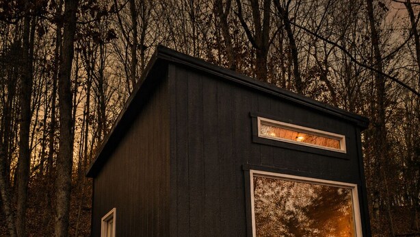 A small, dark wooden cabin with a single window and a door, surrounded by trees.