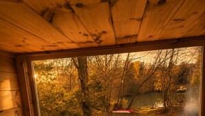 A wooden cabin interior with a bed, a window overlooking a forest and a lake.