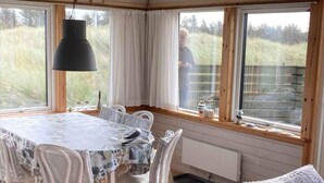 A dining area with a round table, white chairs, a black pendant light, and large windows with curtains. - 4 Star Holiday Home in Lokken (Lokken)