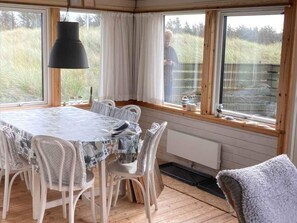 A dining area with a round table, white chairs, a black pendant light, and large windows with curtains. - 4 Star Holiday Home in Lokken (Lokken)