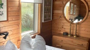 A wooden cabin room with a bed, a dresser, a mirror, and a window with a view of greenery. - 4 Star Holiday Home in Lokken (Lokken)
