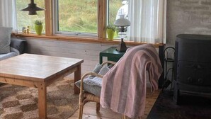 A cozy room with a wooden table, a wicker chair, a sofa, a window with curtains, a lamp, and a wood-burning stove. - 4 Star Holiday Home in Lokken (Lokken)