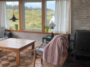 A cozy room with a wooden table, a wicker chair, a sofa, a window with curtains, a lamp, and a wood-burning stove. - 4 Star Holiday Home in Lokken (Lokken)